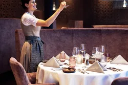 Waitress in traditional dress holding wine glass in upscale restaurant with set dinner table